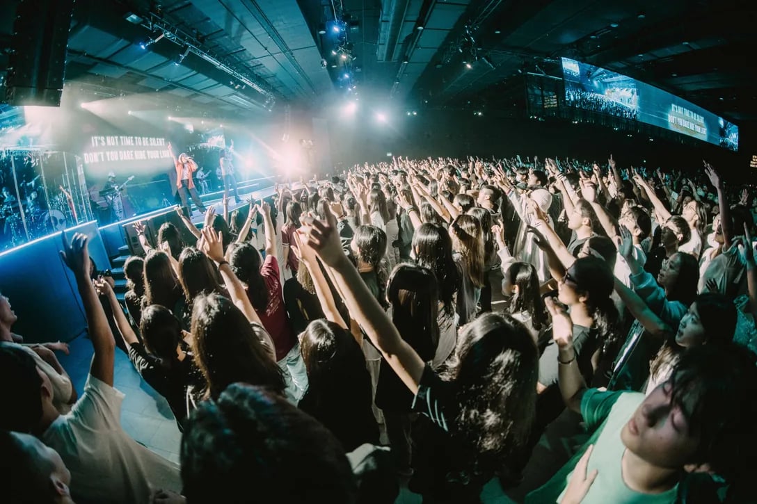 A packed auditorium of young people worshipping with hands raised, stage lights overhead.