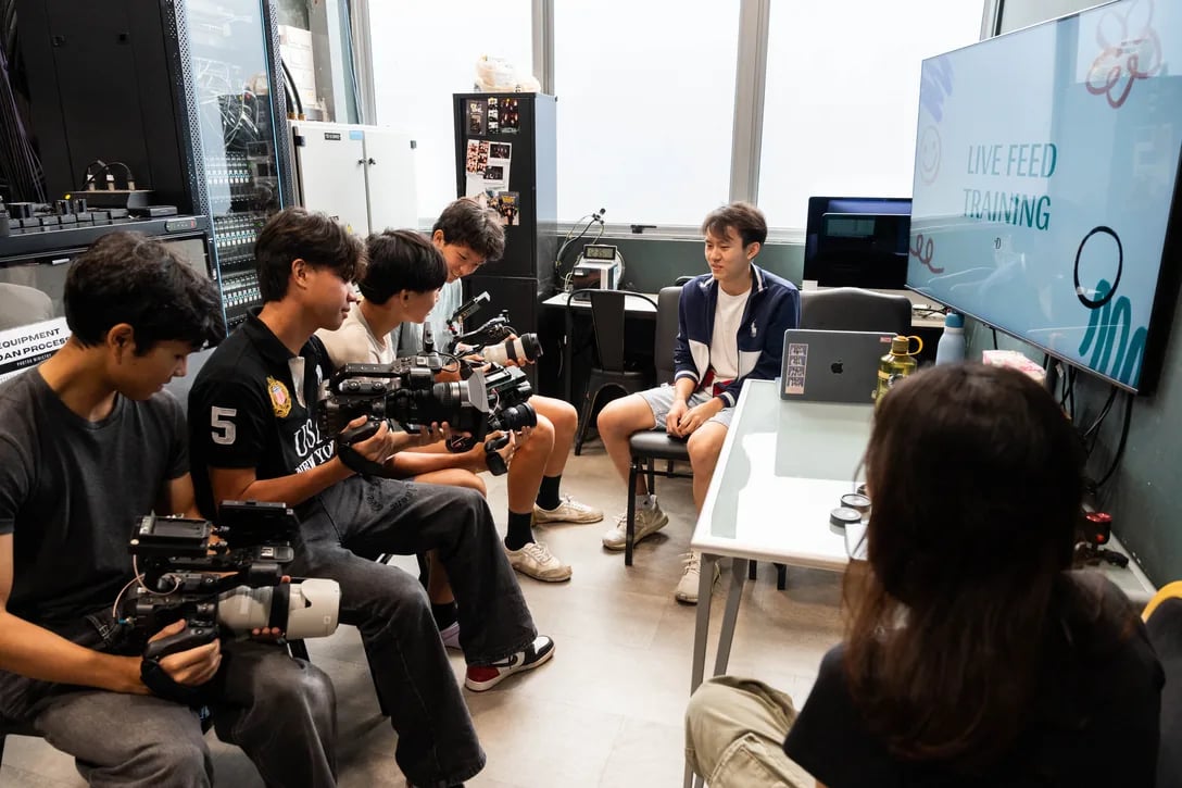 Young people training with professional video cameras in the media room, filming an interview in front of a screen reading Live Feed Training.