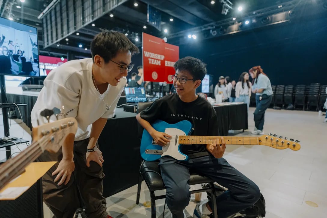 A worship-team member coaching a young person on the electric guitar, a red Worship Team banner behind them.