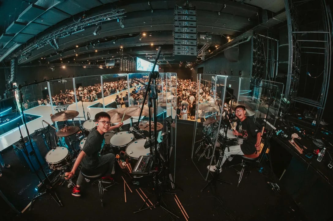 Two young drummers grinning mid-song on stage, a packed auditorium spread out behind them.