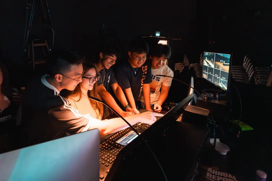 Young people gathered at the sound-mixing console in the tech booth, camera feeds on a monitor beside them.