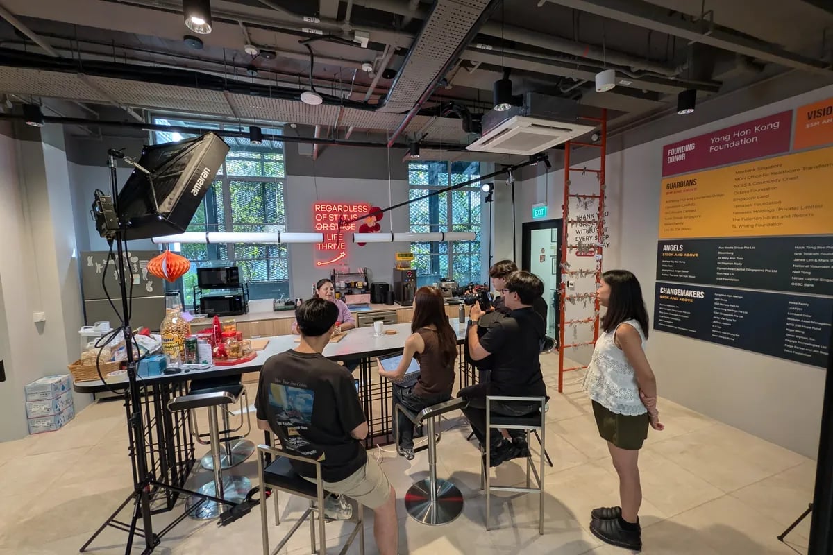 A studio shoot around a high table beneath a neon sign reading 'Regardless of status or stage in life, thrive.'