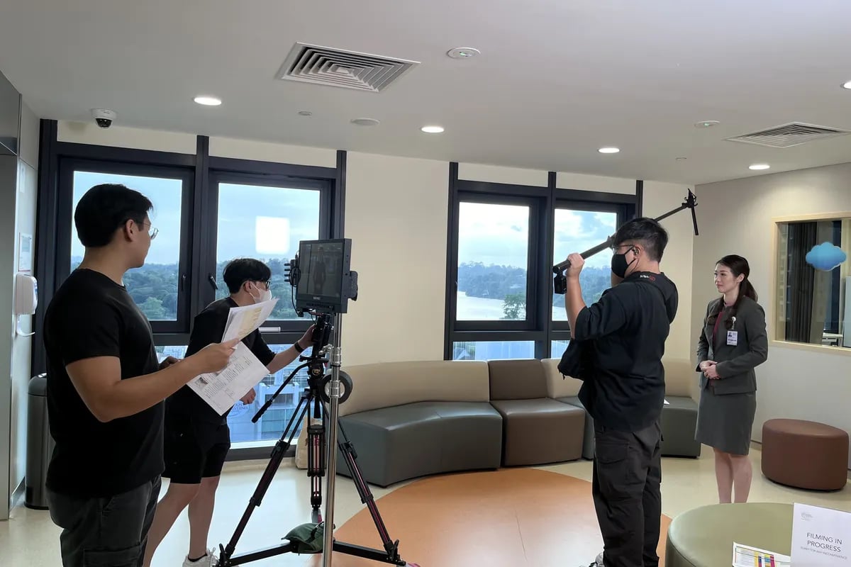 The team filming an interview in a bright lounge, a camera on a tripod facing the sofa.